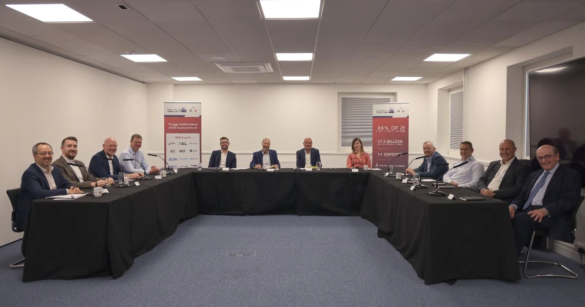 Thirteen people seated around a large U-shaped table in a meeting room with presentation banners in the background