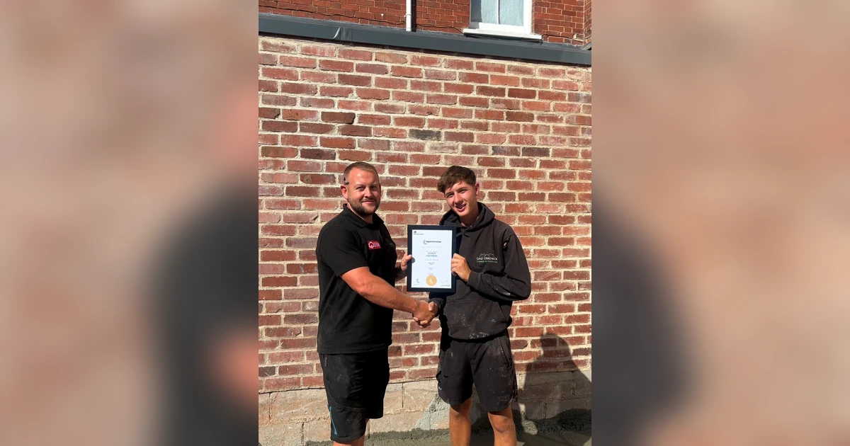 Dale Chadwick shaking hands with apprentice Reuben outside in front of a brick wall, holding an apprenticeship certificate