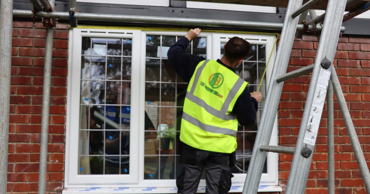 Person wearing hi-vis vest measuring an installed window on a brick house with scaffolding nearby