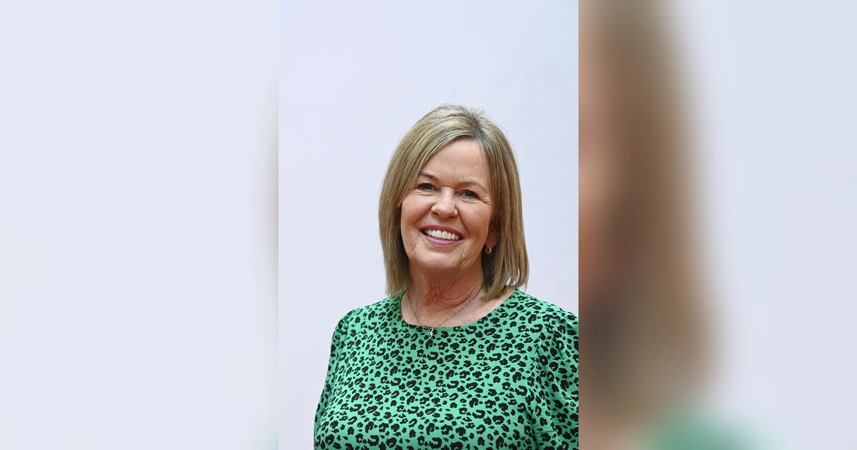 Nickie West smiling, wearing a green patterned top against a plain background