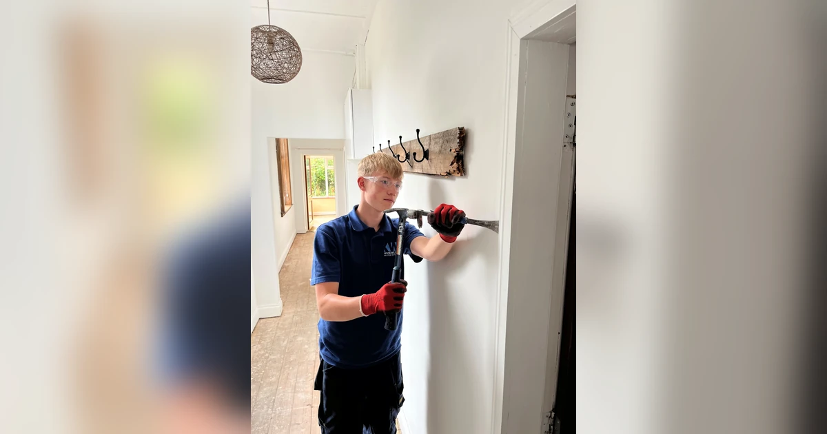 Young apprentice hammering a hook onto a wall inside a building, wearing work gloves and safety glasses