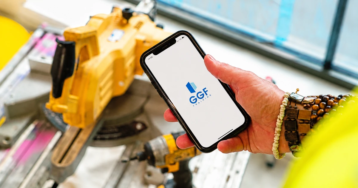 Hand holding smartphone showing GGF Training app, with workshop tools and equipment in the background