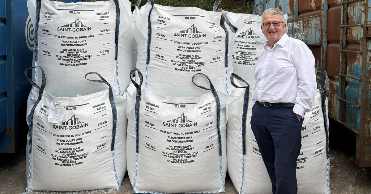 Ian Short, Managing Director of Morley Glass, standing next to stacks of large white bags of recycled glass cullet outdoors.