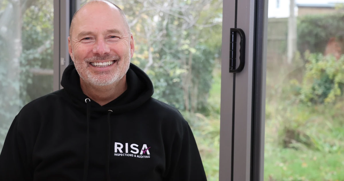 Smiling man wearing black RISA sweatshirt seated indoors with garden visible through window behind him