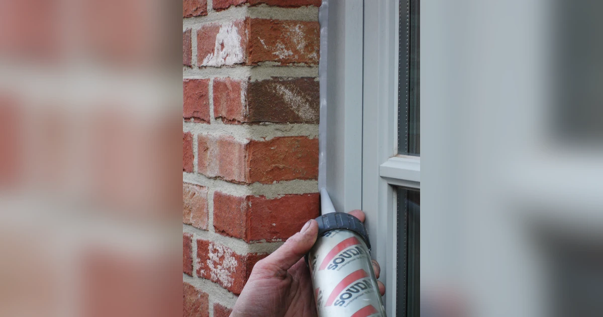 Close-up of a hand applying Soudal sealant to the exterior edge of a window frame set in a brick wall