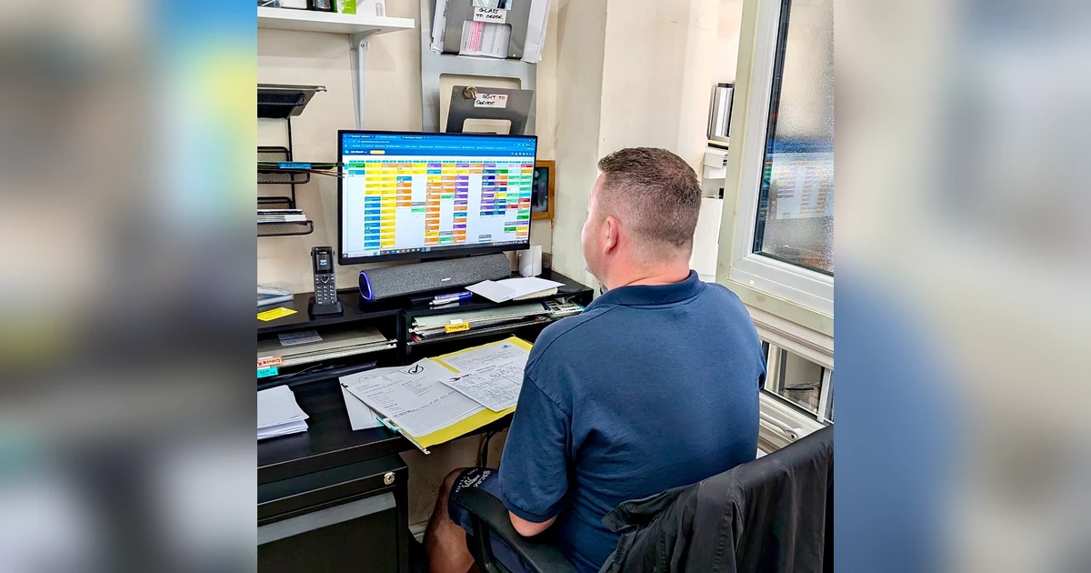 Man seated at a desk working on a computer displaying a colorful job scheduling board with paperwork nearby in an office setting