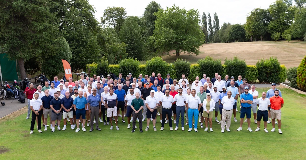 Large group of people standing on a golf course green with trees and golf course landscape in the background at a golf event