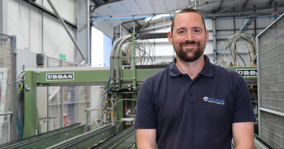 Man in a navy blue Universal Trade Frames polo shirt standing inside a manufacturing facility with machinery in the background