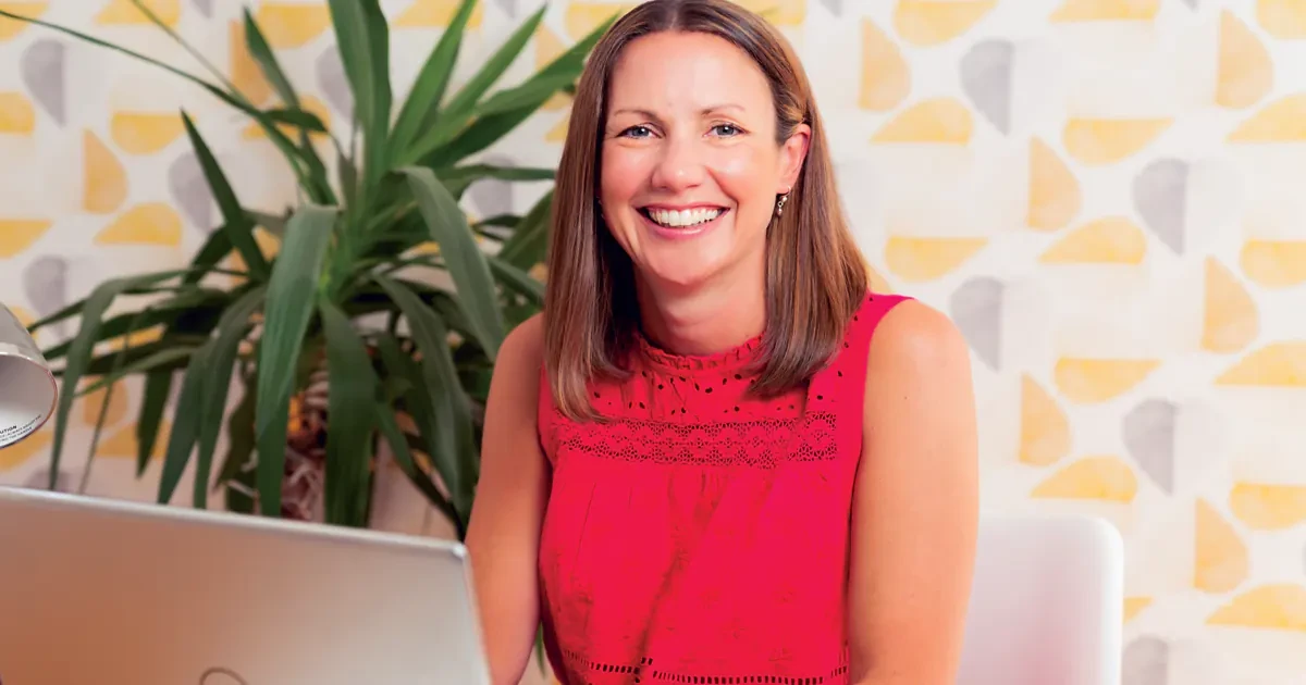 Smiling woman in a red sleeveless top sitting at a desk with a laptop and plant in the background