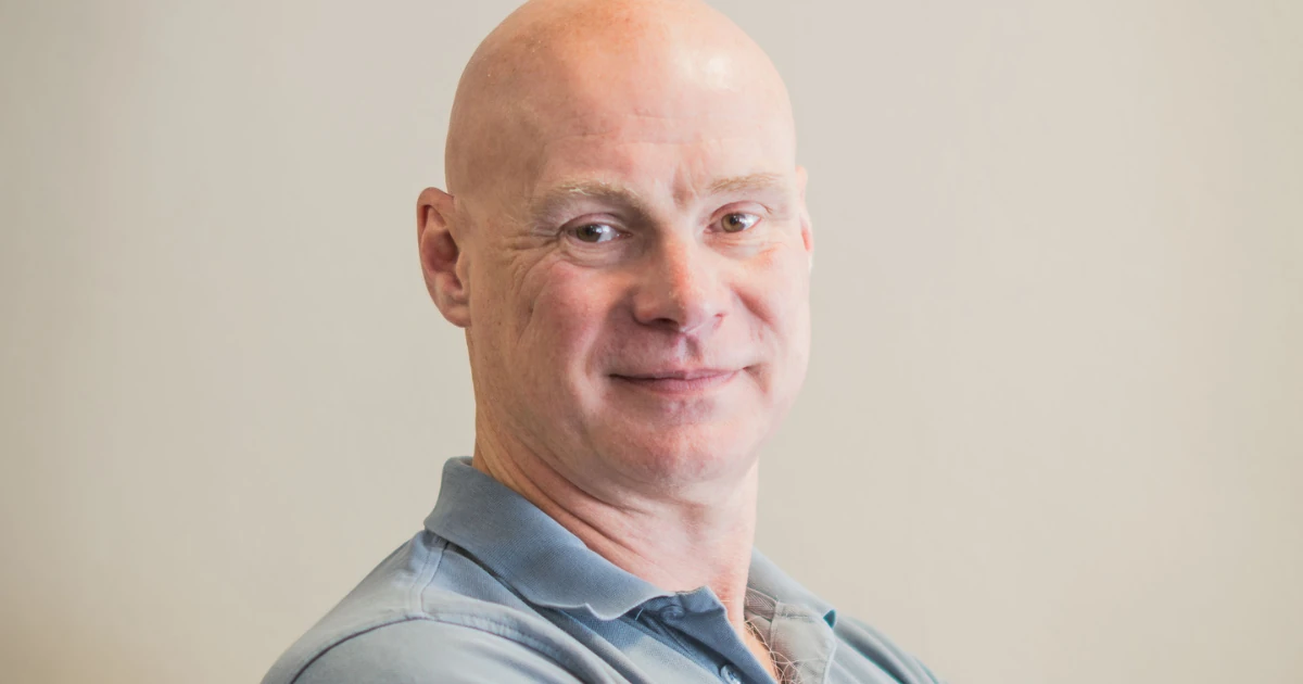 Simon Jarman standing indoors wearing a light blue polo shirt with folded arms