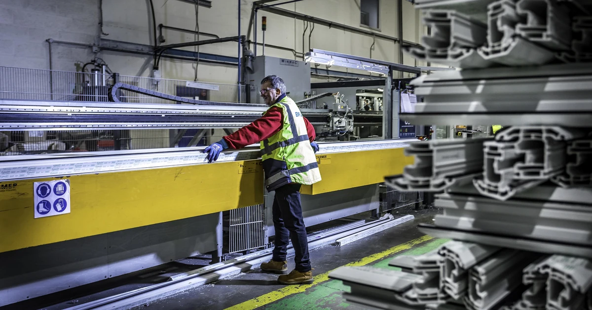Factory worker in high-visibility vest handling window frame machinery surrounded by production materials