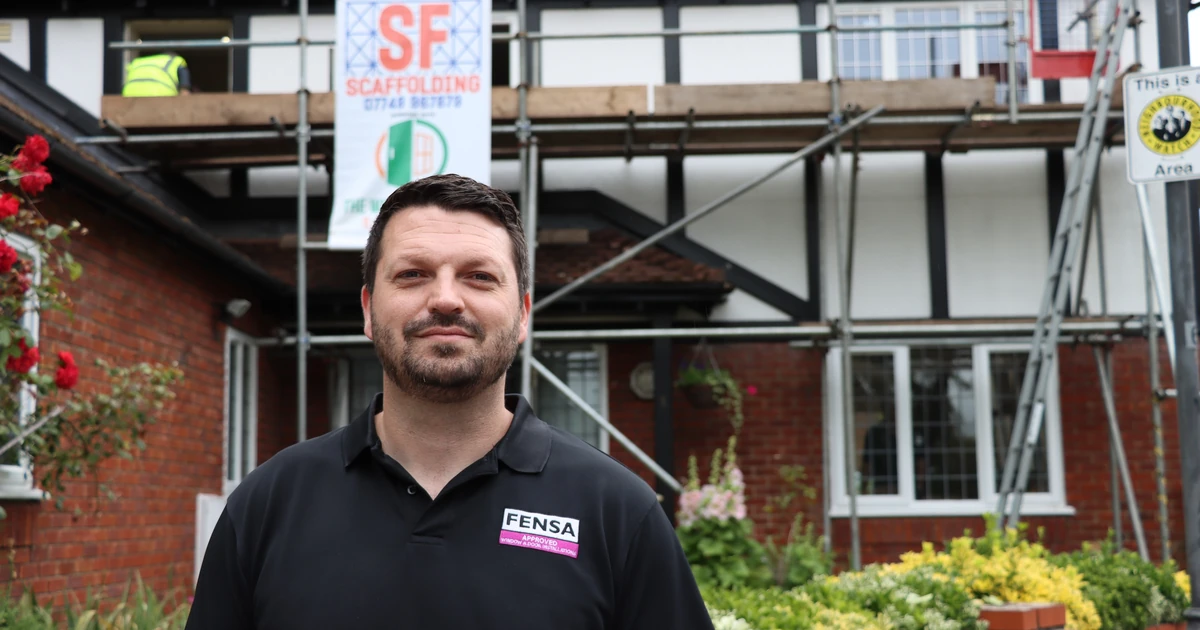 Man standing in front of a house with scaffold, wearing a black FENSA polo shirt, symbolizing professional window installation advice.