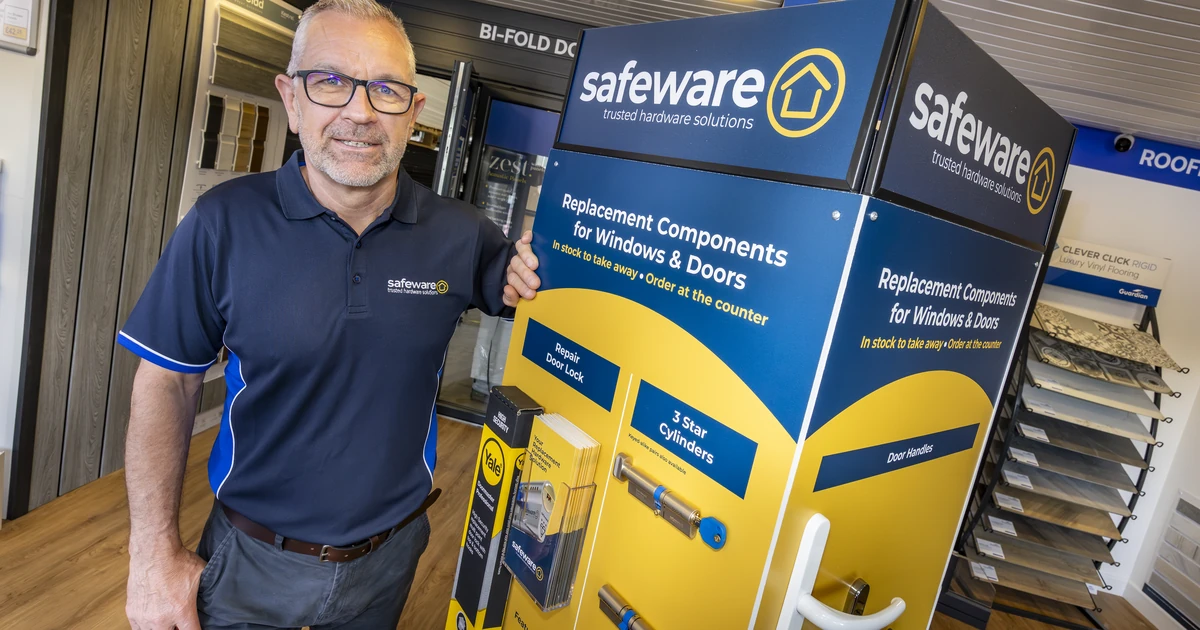 Man in Safeware shirt standing beside a Safeware branded carousel display with replacement window and door hardware in a trade counter setting