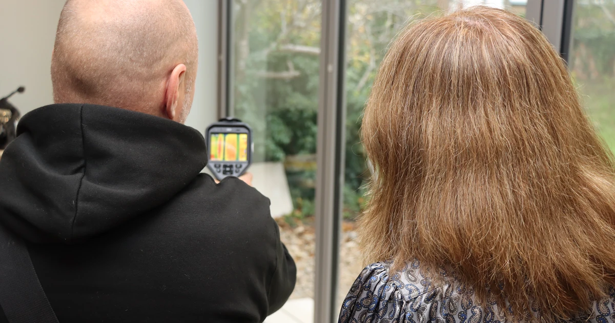 Two people viewed from behind inspecting a large glazed window with a thermal imaging device, highlighting heat performance concerns in glazing