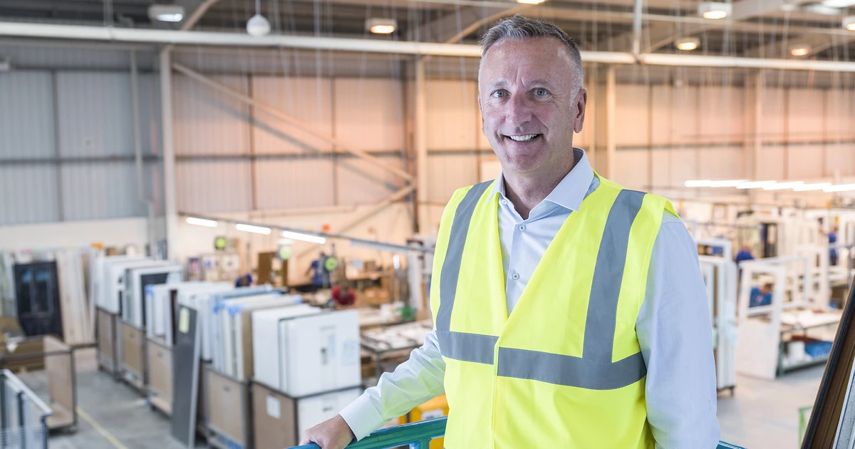 Smiling man wearing a high-visibility vest standing inside a production facility with window and door panels in the background