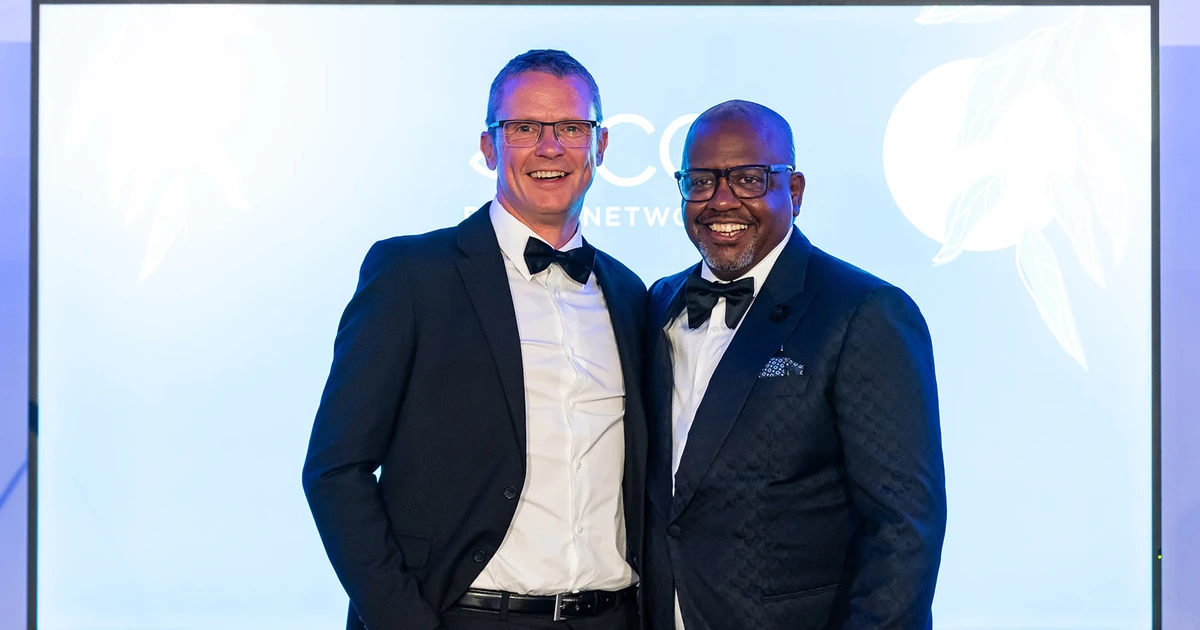 Two men in tuxedos smiling, standing in front of a screen at an event, presumably Greg Kane and Mick Giscombe