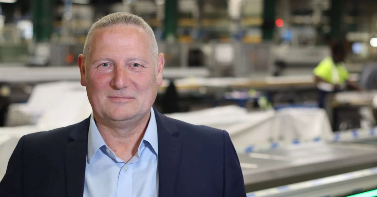 Man in a business suit smiling in an industrial manufacturing setting with workers and machinery in the background
