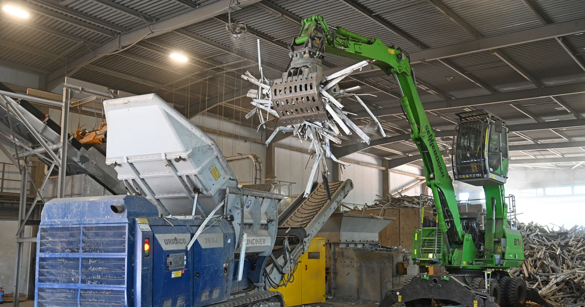 Green mechanical arm loading PVC-U window frames into blue recycling machinery inside a processing facility