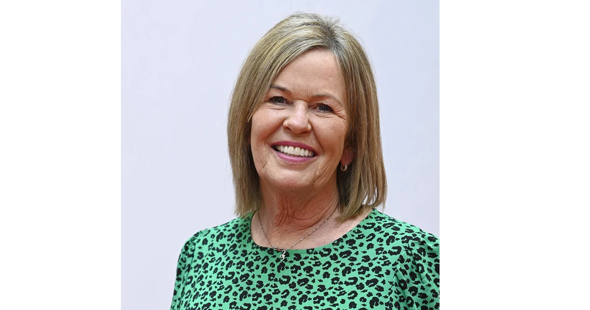 Nickie West smiling in a green patterned top against a plain background