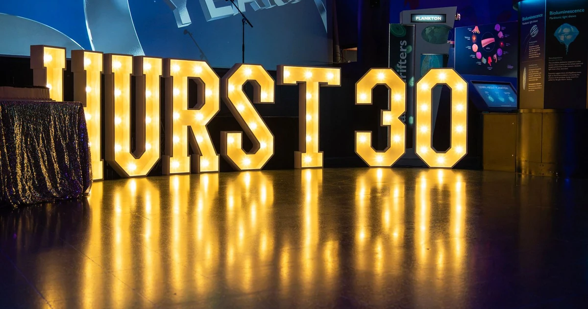 Illuminated letters spelling 'HURST 30' reflecting on a shiny floor at an event venue