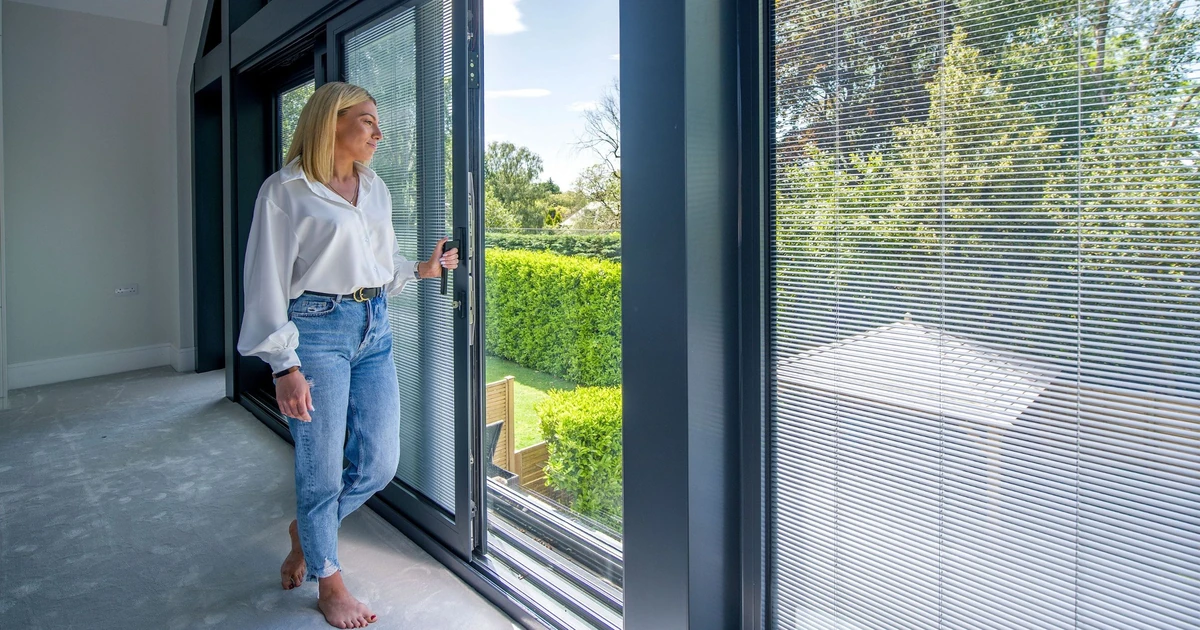 Woman standing inside near a large glass door with integrated blinds looking outside into a garden