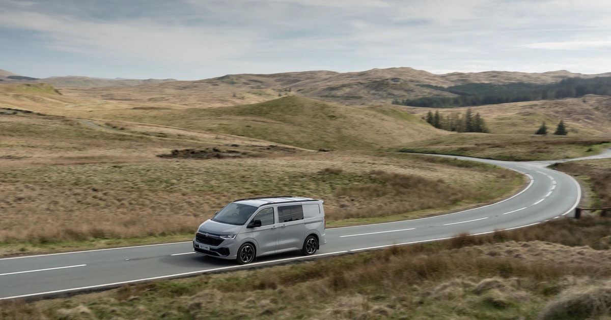 Grey Volkswagen Transporter van driving on a winding rural road with hills in the background